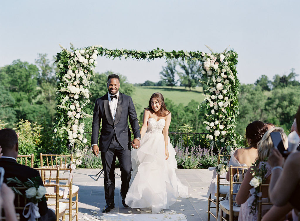 A couple walks down the aisle with a floral arch behind them at an outdoor ceremony at Shadow Creek wedding venue in Virginia