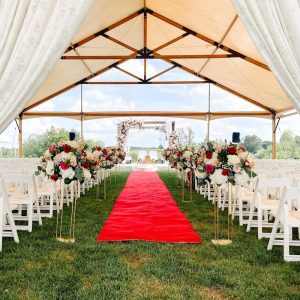 A red carpet stretches down the aisle at an outdoor tented wedding ceremony surrounded by red and white flowers before guests arrive at Shadow Creek venue in Virginia