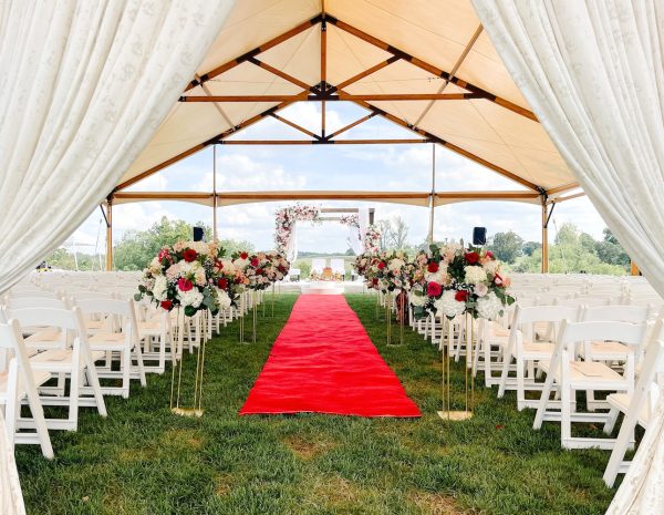 A red carpet stretches down the aisle at an outdoor tented wedding ceremony surrounded by red and white flowers before guests arrive at Shadow Creek venue in Virginia