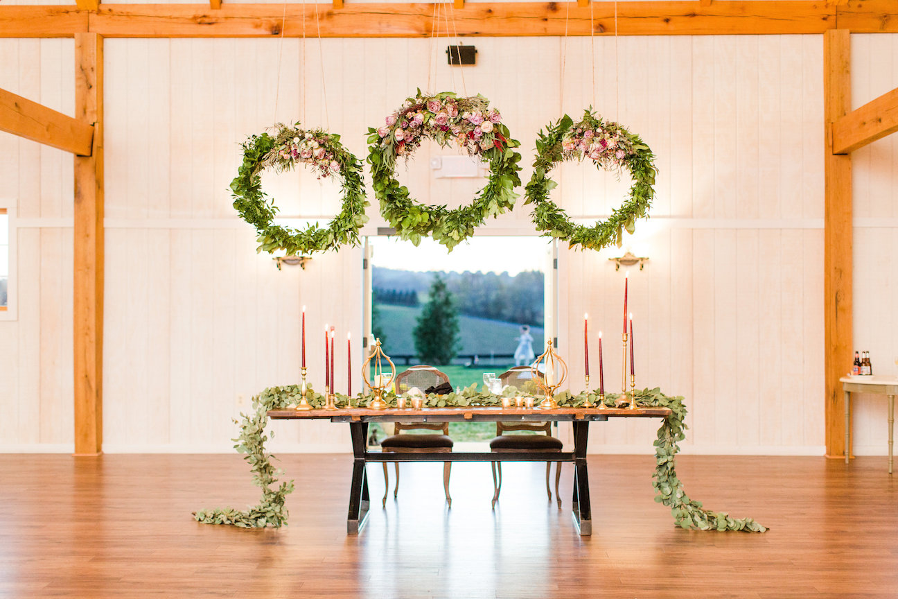 A sweetheart table covered in gorgeous flowers with three floral wreaths hanging above it inside at Shadow Creek wedding barn in Virginia
