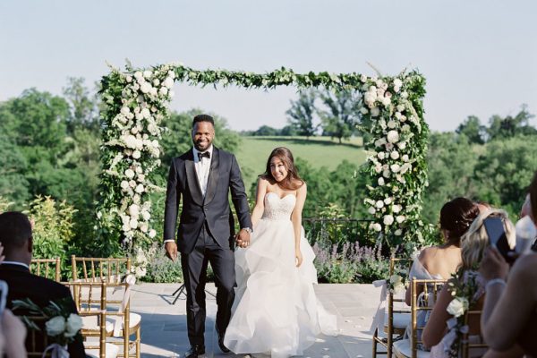 A couple walks down the aisle with a floral arch behind them at an outdoor ceremony at Shadow Creek wedding venue in Virginia